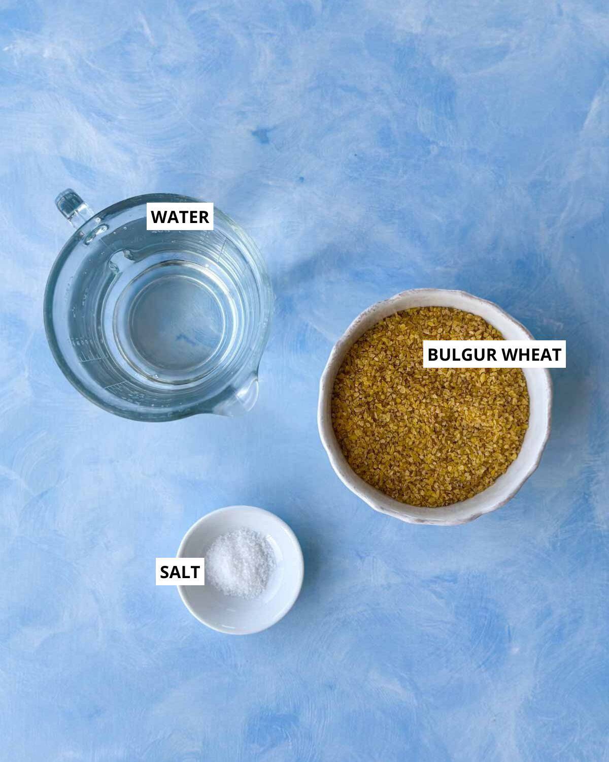Labeled overhead photo showing ingredients for cooking bulgur wheat on the stovetop: a jug of water, a bowl of coarse bulgur wheat, and a small dish of salt on a light blue background.