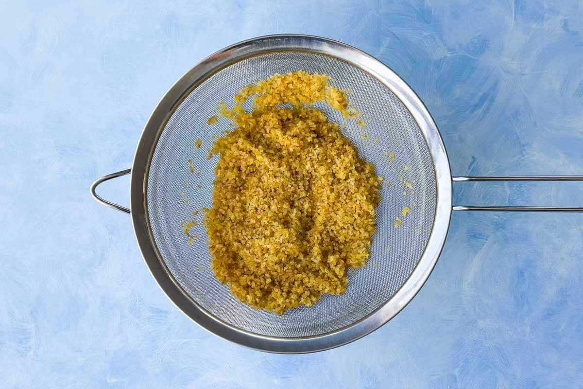 Drained cooked bulgur wheat resting in a fine mesh sieve over a blue background.