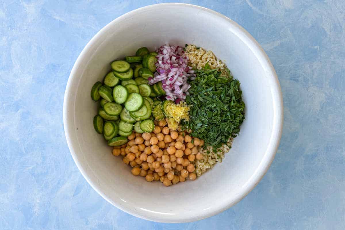Salad ingredients arranged in a large bowl, including chopped parsley, red onion, cucumber, lemon zest, chickpeas, and bulgur wheat.
