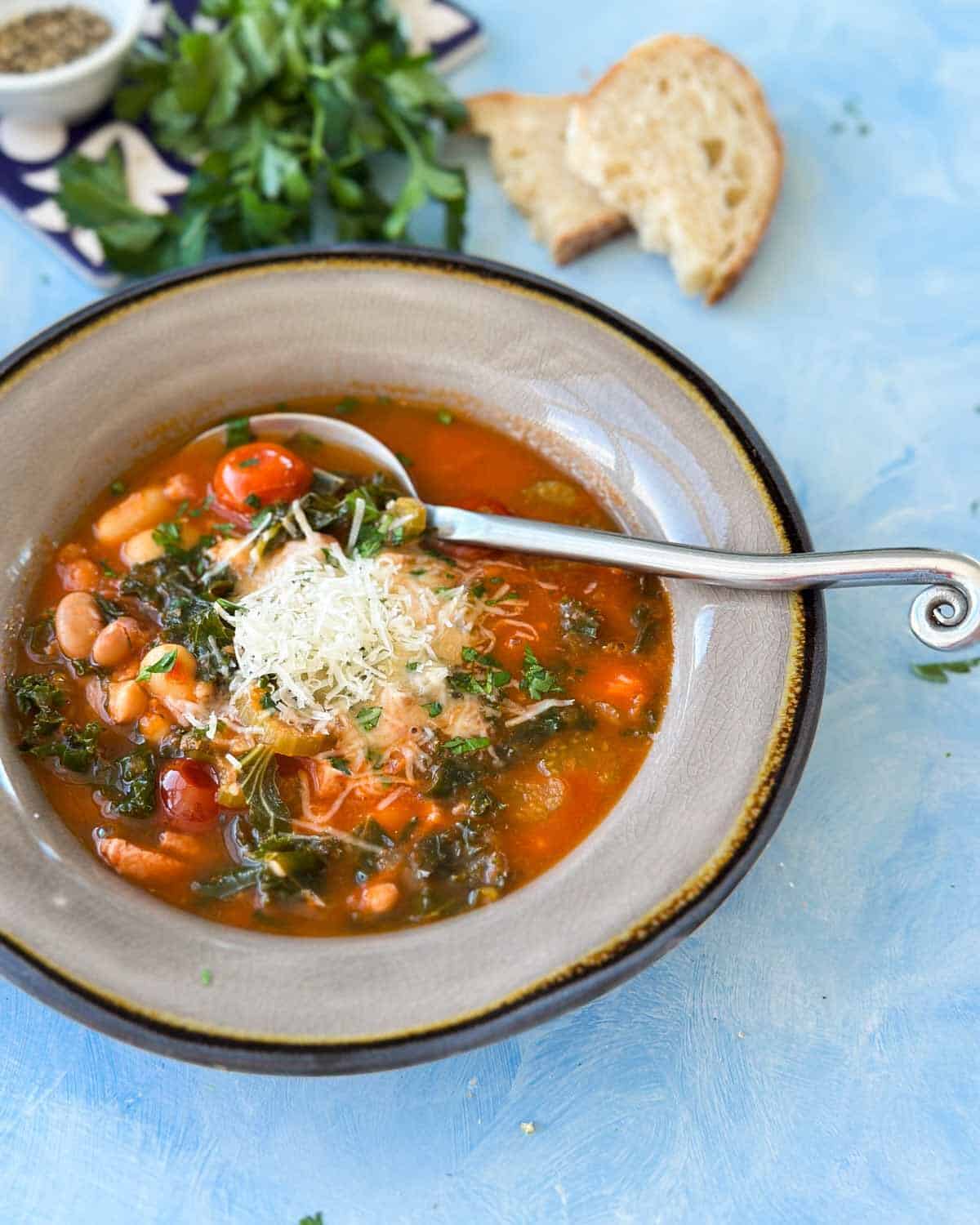 Angled shot of two bowls of Italian Bean Soup, garnished with Parmesan and parsley, served with bread slices and fresh herbs.