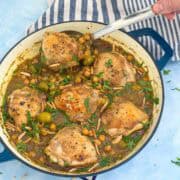 Blue Dutch oven filled with Moroccan chicken tagine, showing chicken thighs, chickpeas, green olives, preserved lemon, almonds, and parsley with a serving spoon.
