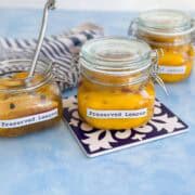Trio of glass jars filled with preserved lemons on a patterned tile and blue surface, one open with a spoon and striped towel in the background.