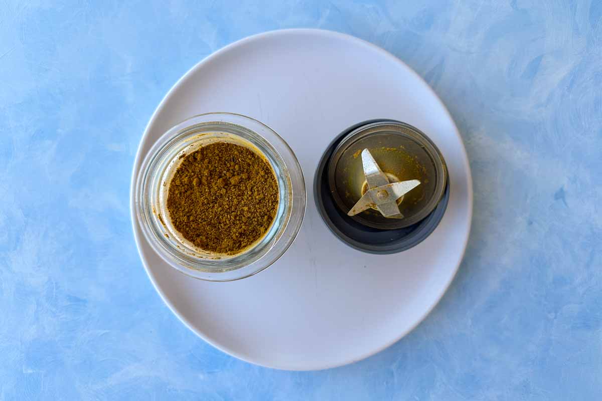 Overhead view of freshly ground Moroccan tagine spice blend in a glass jar, placed beside a spice grinder on a white plate with a blue background.