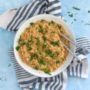 Overhead view of a white bowl filled with fluffy toasted couscous, garnished with fresh parsley. A striped towel rests underneath on a blue background. Great example of how to toast couscous at home.