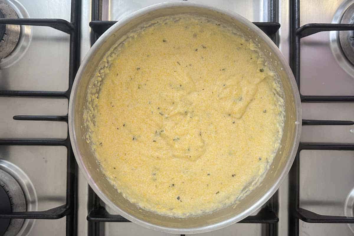 Creamy polenta cooking in a saucepan until thick and smooth with flecks of rosemary throughout.