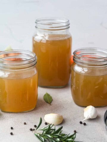 Close-up of three jars of golden beef stock with garlic, rosemary, peppercorns, and herbs on the countertop.