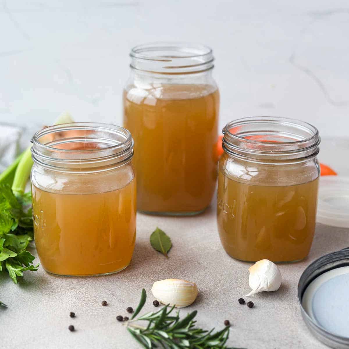 Close-up of three jars of golden beef stock with garlic, rosemary, peppercorns, and herbs on the countertop.