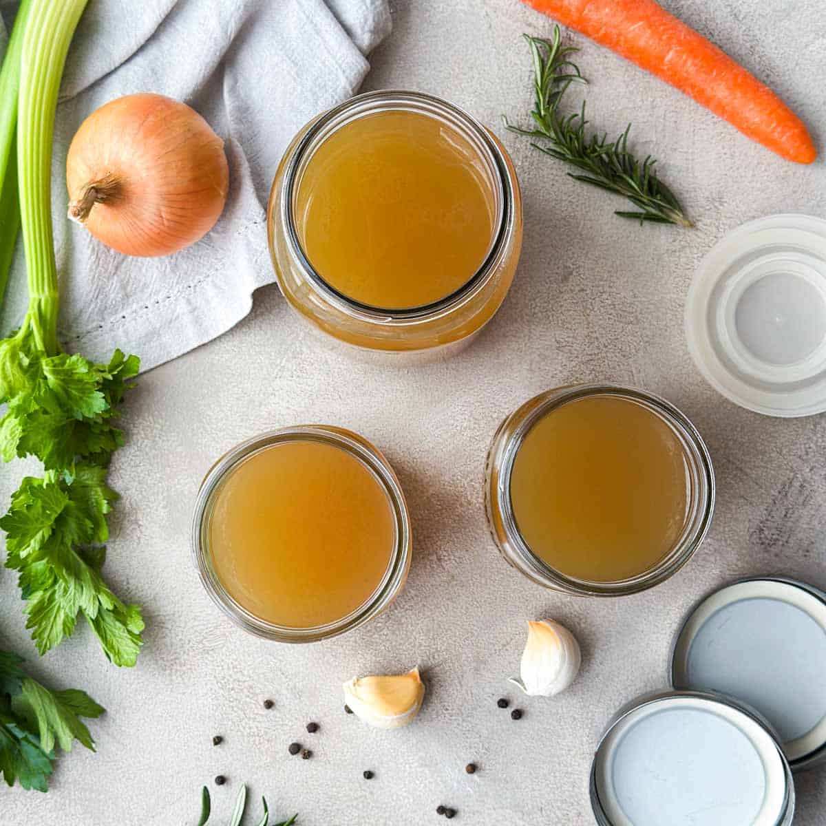 Overhead view of three jars of homemade beef stock surrounded by celery, carrot, onion, garlic, rosemary, and peppercorns.