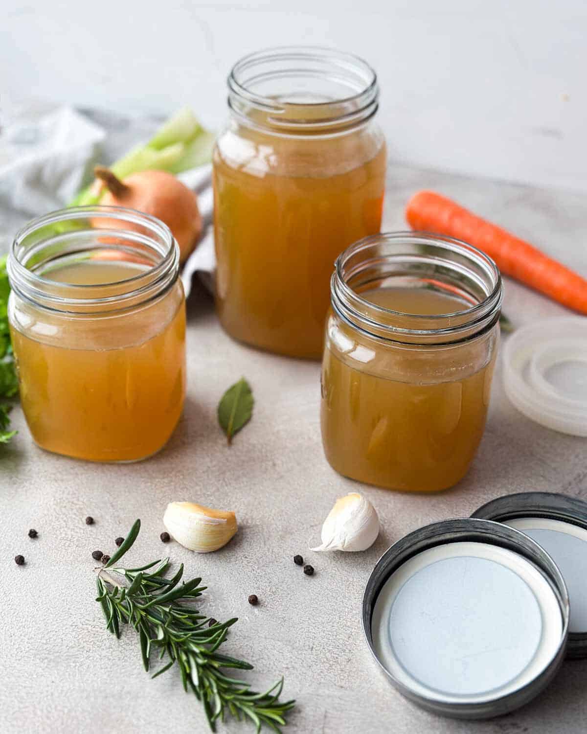 Portrait close-up of glass jars filled with homemade beef stock, styled with garlic, rosemary, peppercorns, and lids.