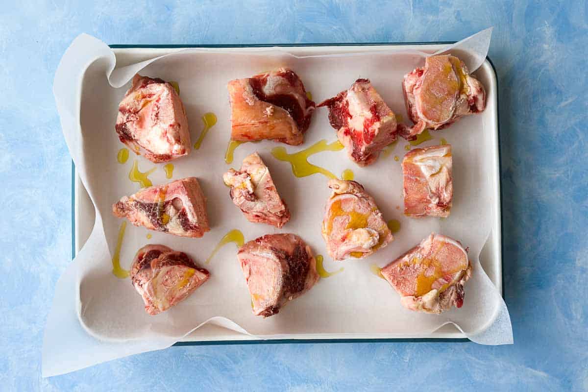 Top view of raw beef bones drizzled with olive oil on a parchment-lined baking tray before roasting for homemade beef stock.