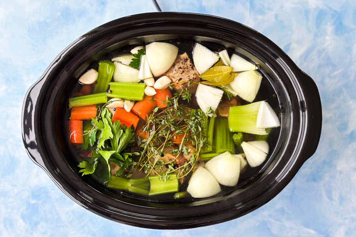 Top view of a slow cooker filled with roasted beef bones, celery, carrots, onion, garlic, herbs, bay leaves, and water for beef stock.
