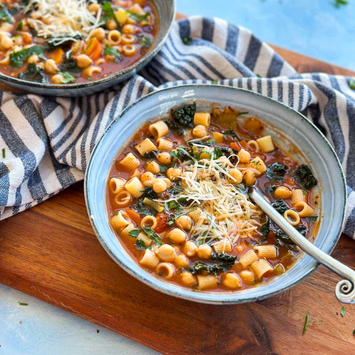 Two bowls of pasta e ceci soup with chickpeas, vegetables, and fresh herbs, served on a wooden board with striped napkins.