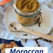 Rustic setup of a Moroccan spice blend in a clip-top jar with whole spices and a wooden spoon on a linen napkin. Text overlay “DIY Moroccan Spice Blend – 2 Easy Ways!” for an inviting and informative visual.