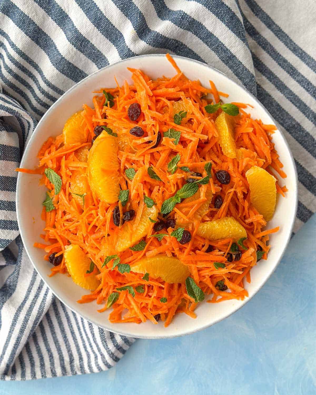 Overhead view of Moroccan carrot salad with oranges, raisins, and fresh mint on a striped cloth.
