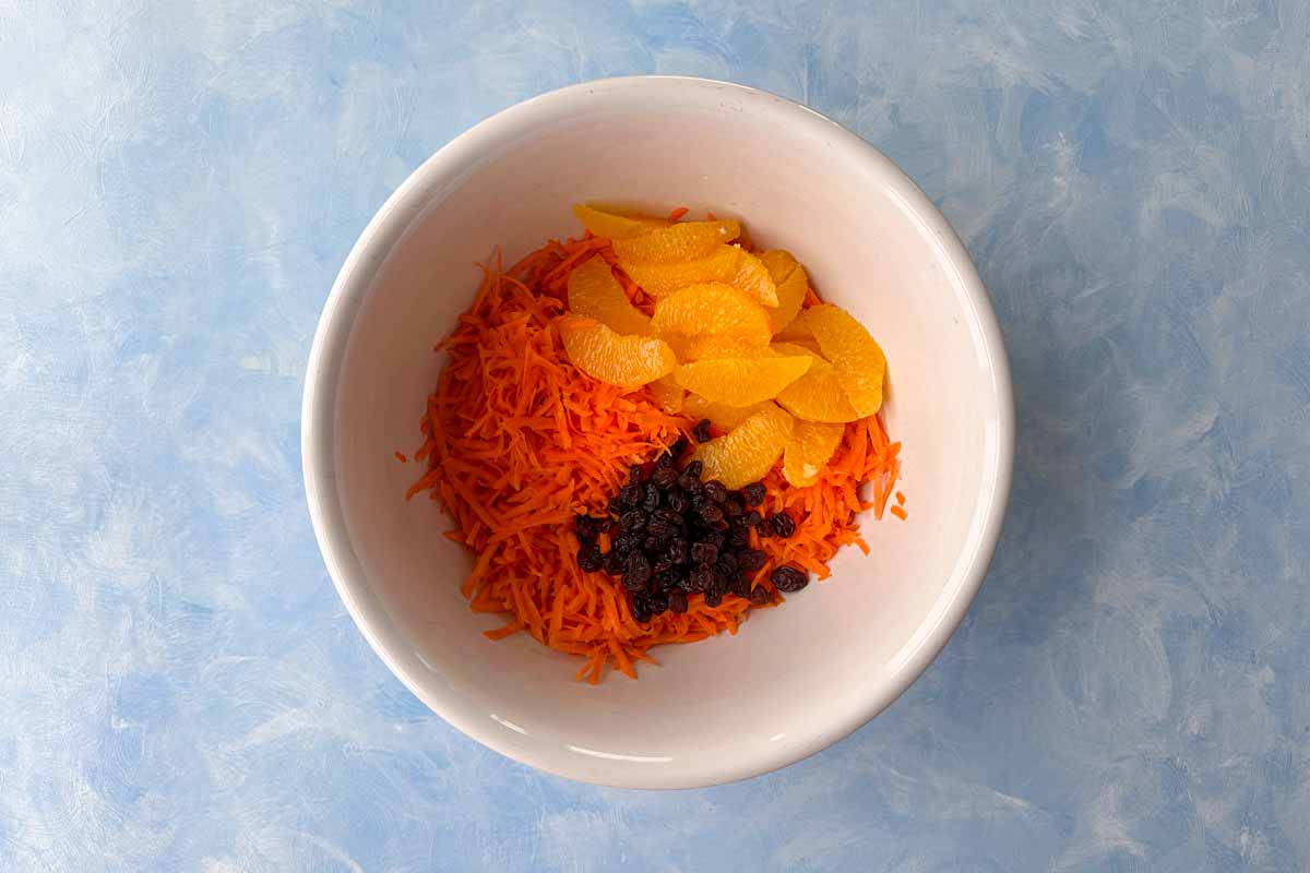 Shredded carrots with fresh orange slices and raisins in a white mixing bowl, ready to be combined for Moroccan carrot salad.