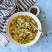 Overhead bowl of chicken orzo soup with shredded chicken, carrots, and herbs, served with a striped napkin and black pepper on a light blue background.