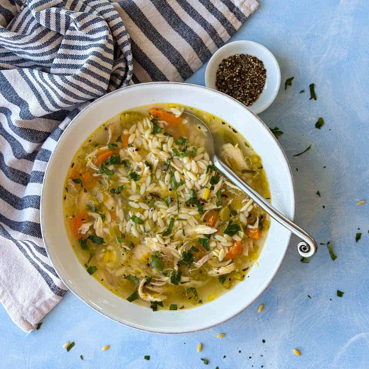 Overhead bowl of chicken orzo soup with shredded chicken, carrots, and herbs, served with a striped napkin and black pepper on a light blue background.