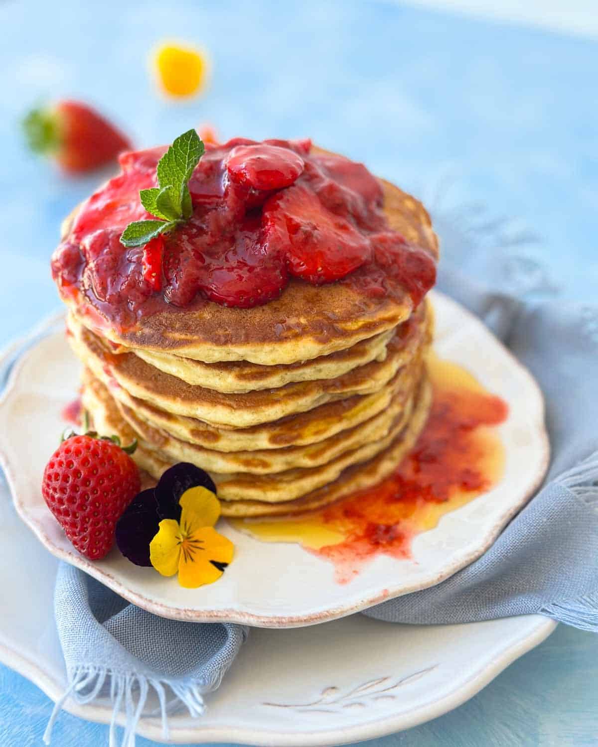 Fluffy Greek yogurt pancakes stacked high and topped with strawberry compote, honey drizzle, fresh strawberries, and edible flowers on a soft blue background.