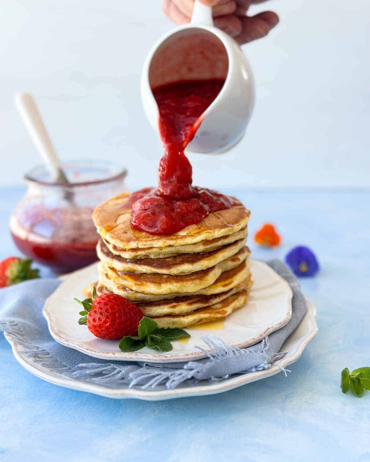 Close-up of strawberry compote being poured over warm Greek yogurt pancakes, highlighting the glossy texture and vibrant color.