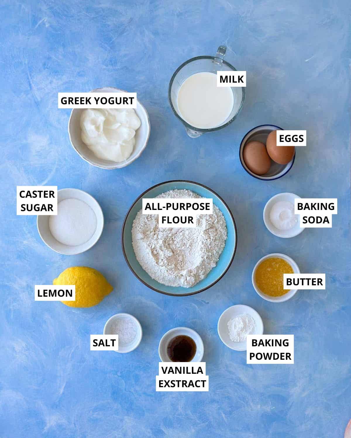 Overhead view of measured ingredients including Greek yogurt, flour, eggs, milk, butter, sugar, lemon, and vanilla extract arranged neatly on a blue surface.