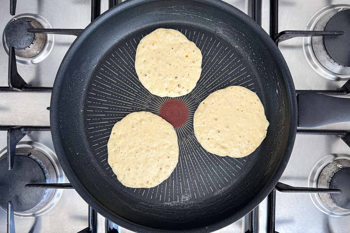 Three pancakes cooking in a non-stick pan, with bubbles forming on the surface as they begin to set.