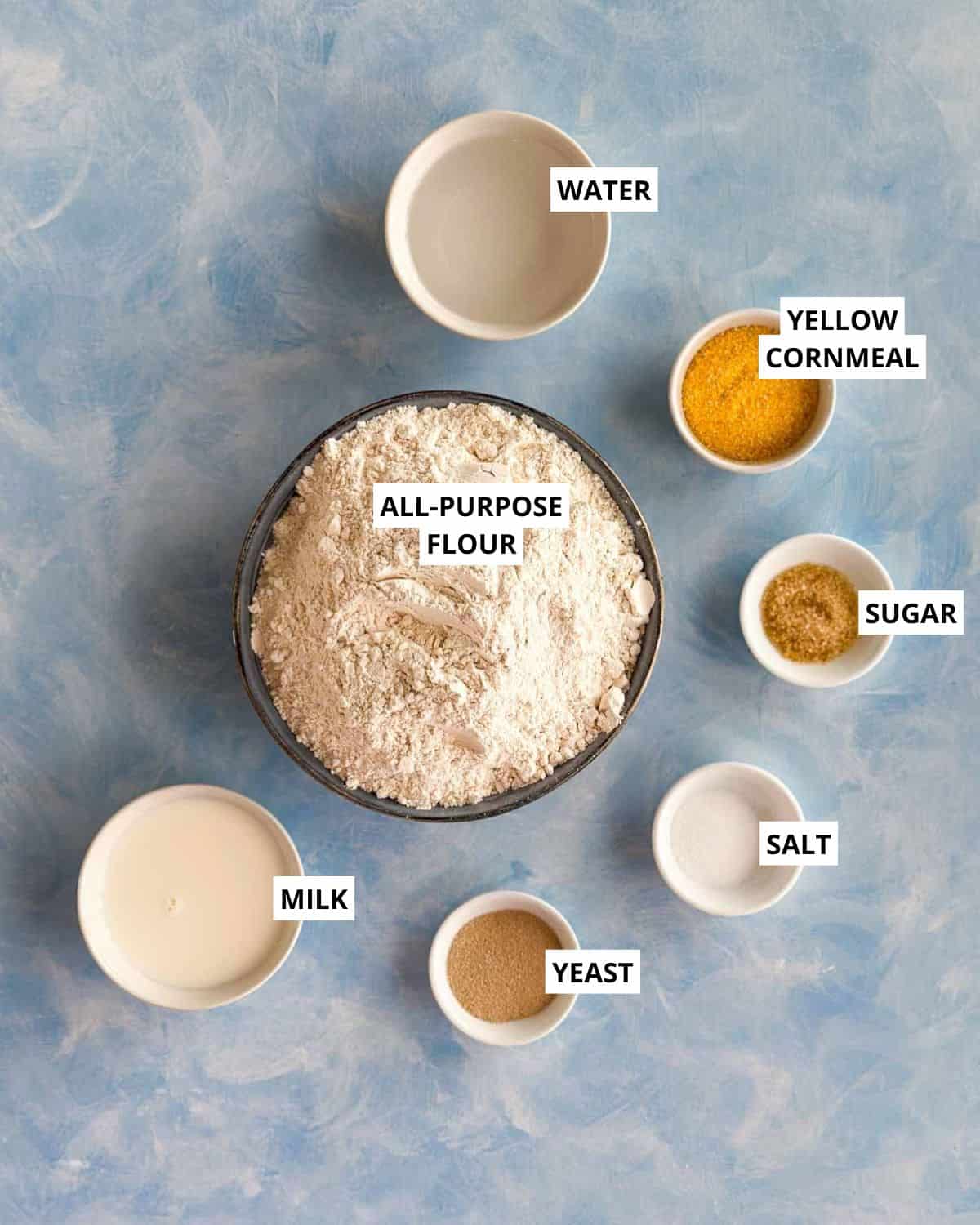 Overhead shot of ingredients for Moroccan bread (khobz kesra) including flour, cornmeal, milk, yeast, saffron, sugar, salt, and water arranged on a blue background.