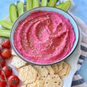 Top-down view of vibrant pink roasted beet hummus surrounded by seeded crackers, fresh cherry tomatoes on the vine, and sliced cucumbers.