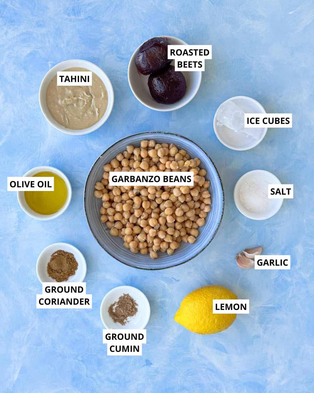 Ingredients for roasted beet hummus including garbanzo beans, roasted beets, tahini, olive oil, garlic, lemon, ground cumin, ground coriander, salt, and ice cubes arranged on a blue background.