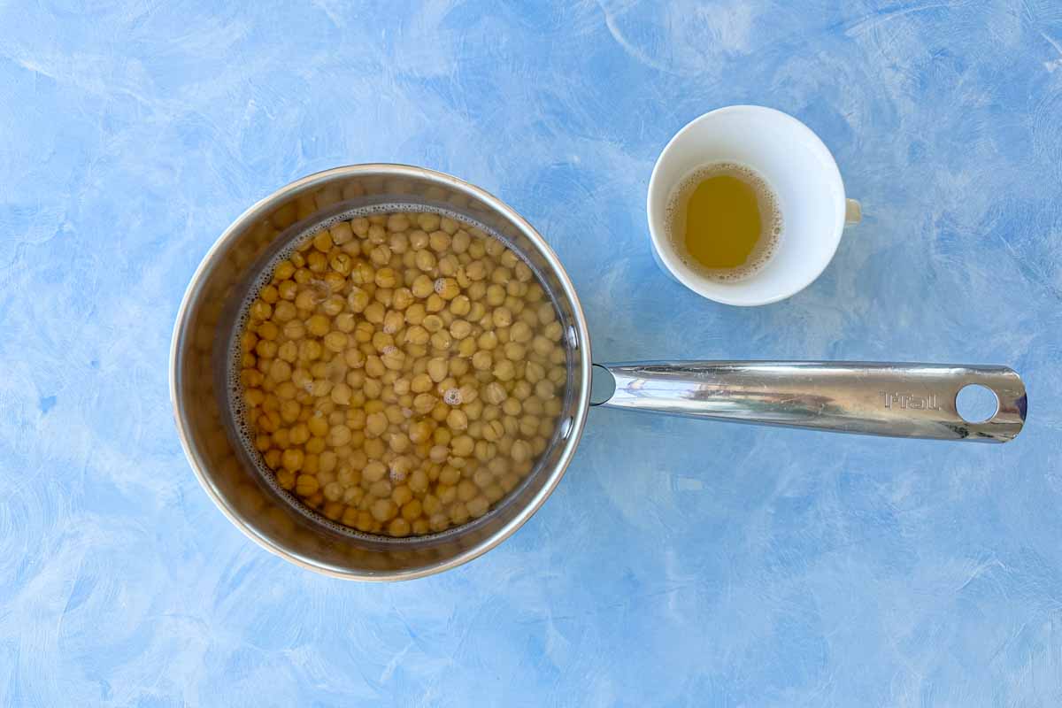 Cooked garbanzo beans with reserved chickpea cooking liquid (aquafaba) shown in bowls on a blue background for making homemade hummus.