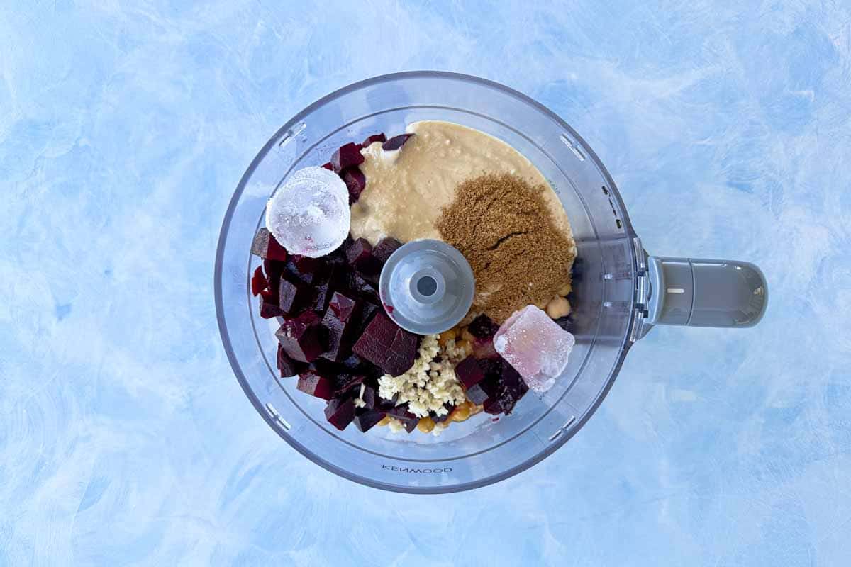Roasted beets, garbanzo beans, tahini, garlic, lemon juice, spices, olive oil, ice cubes, and aquafaba added to a food processor before blending roasted beet hummus.