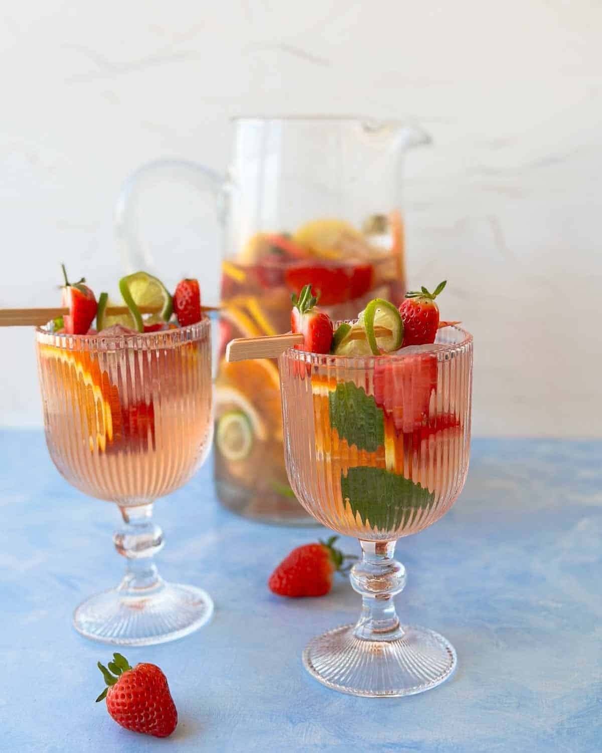 Summer rosé sangria with fresh strawberries, citrus slices, and mint, served over ice with a glass pitcher of sangria behind.