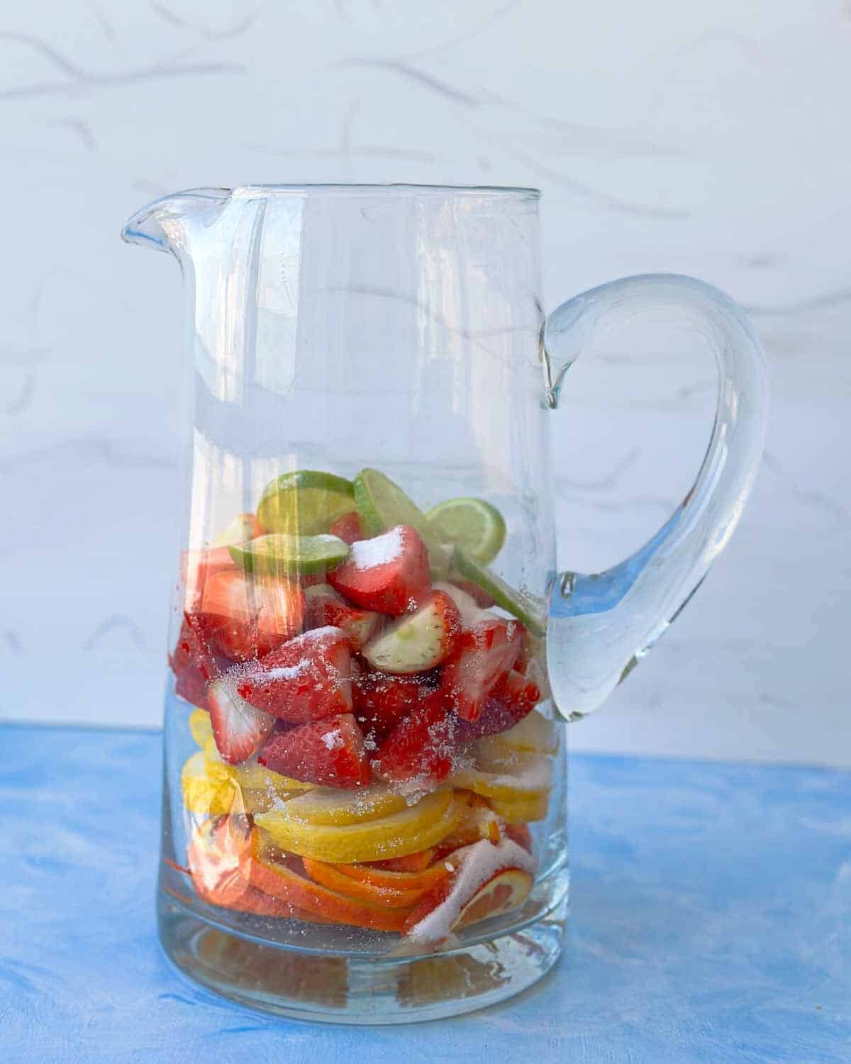 Fresh strawberries and sliced citrus layered in a glass pitcher with caster sugar, ready to soak and infuse for homemade rosé sangria.