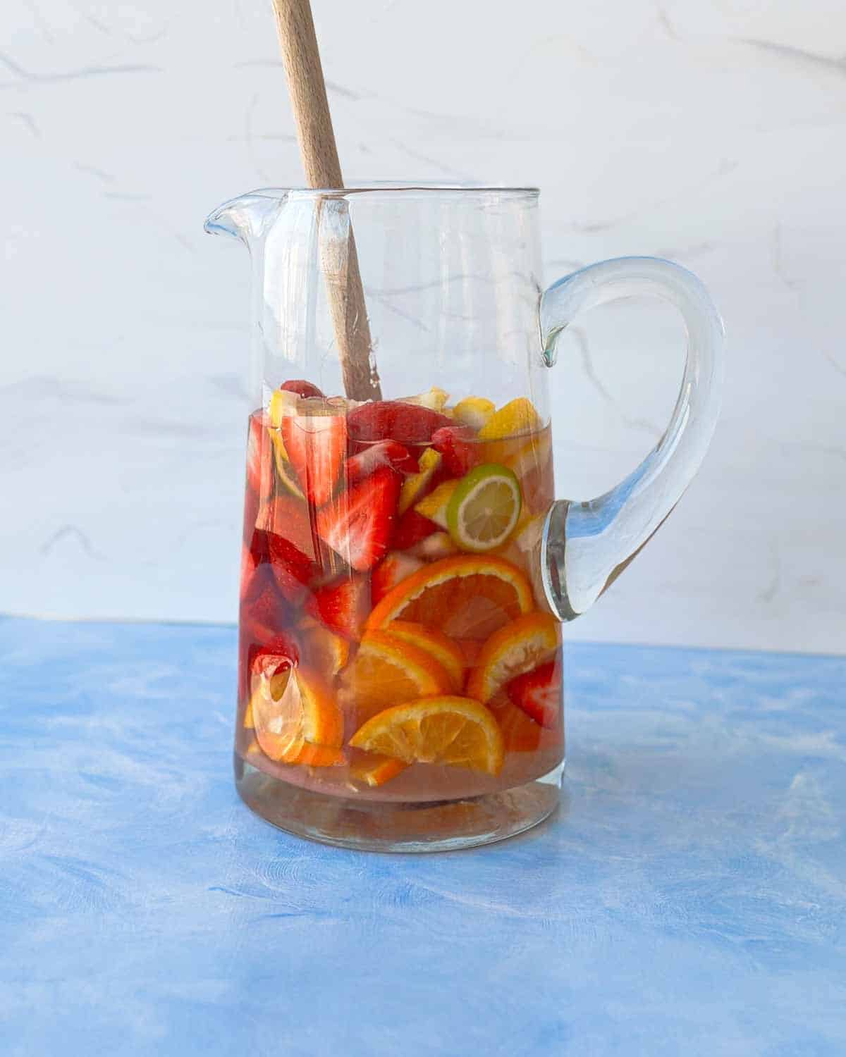 Rosé wine poured over strawberries and citrus in a glass pitcher, gently stirred with a wooden spoon as the sangria begins to come together.