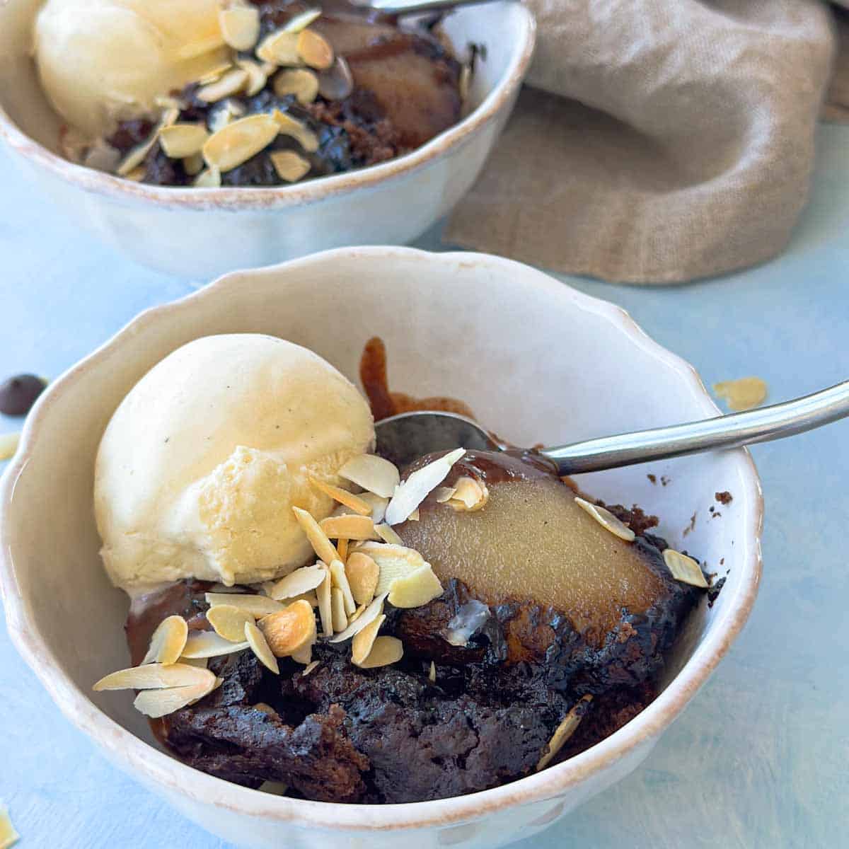 Bowl of self-saucing chocolate pudding served with a poached pear, vanilla ice cream, and scattered almonds, showing the molten chocolate sauce.
