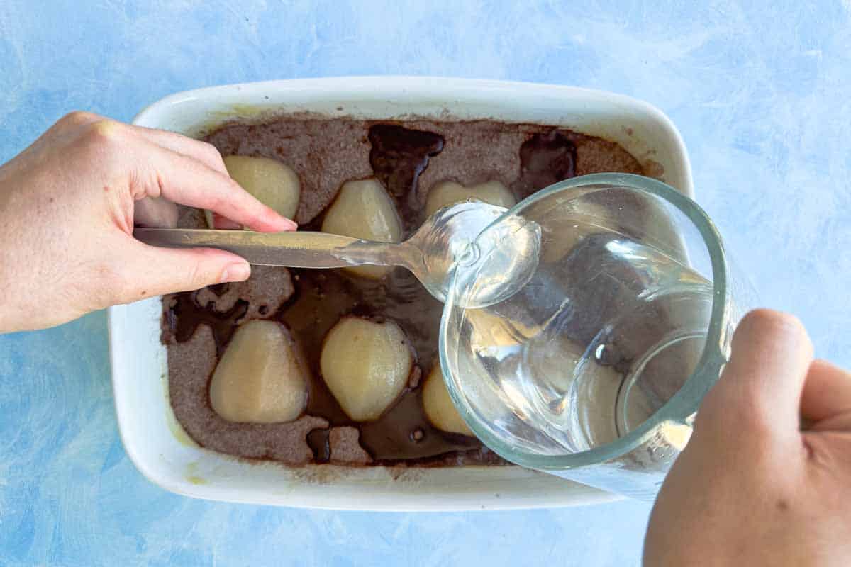 Water being poured over the back of a spoon into the chocolate pudding dish to create the self-saucing layer, with pears visible beneath.