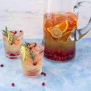 Pitcher of white wine sangria with citrus slices, lime, and pomegranate seeds beside two glasses garnished with rosemary and lime on a blue background.