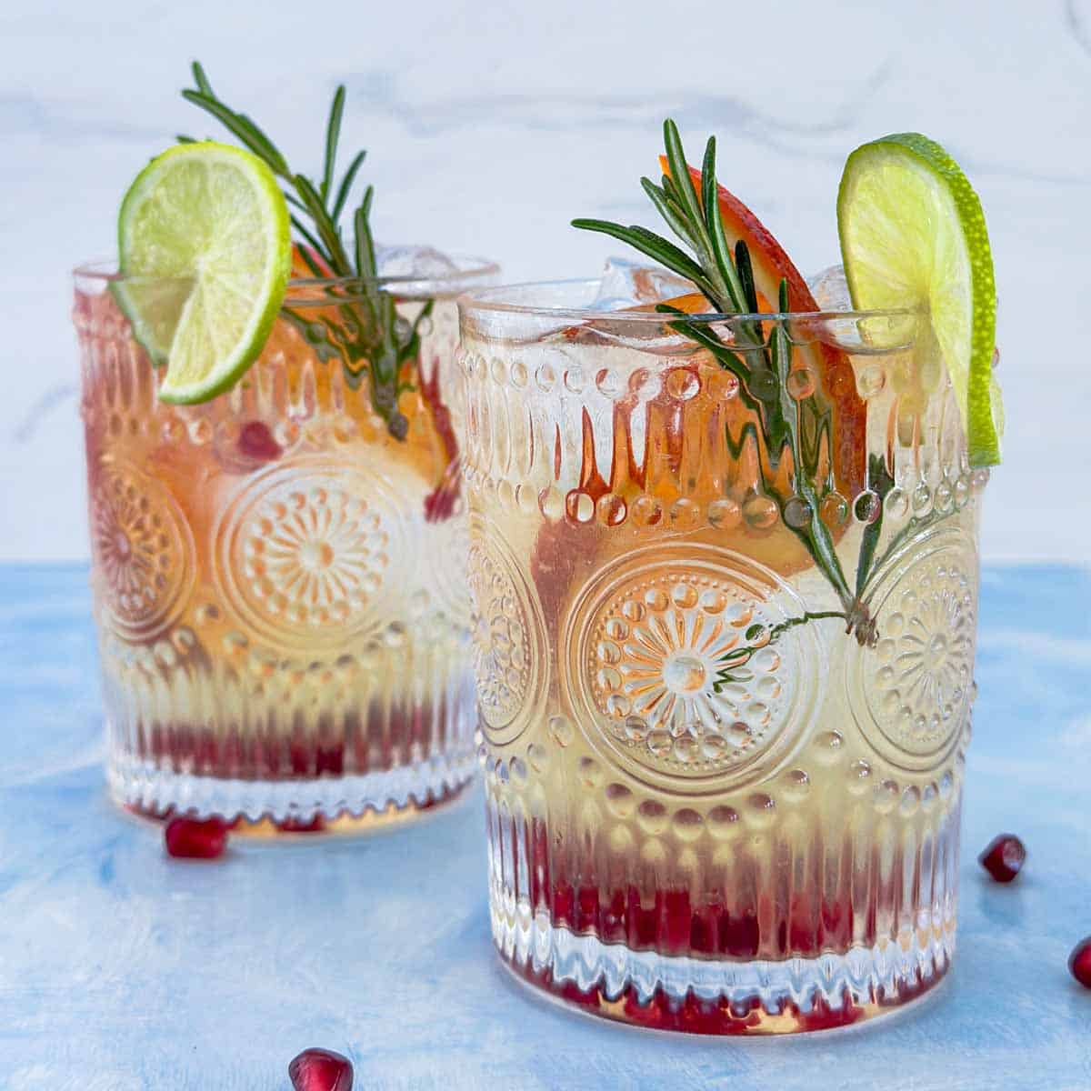 Close-up of two glasses of white wine sangria with orange slices, rosemary sprigs, and lime wedges over ice.