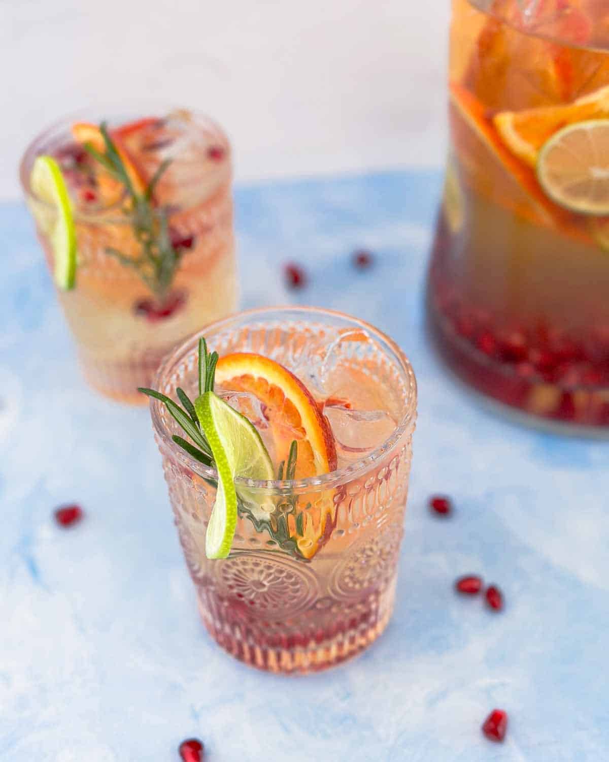 Angled shot of glasses of white wine sangria with citrus and rosemary garnish beside a pitcher filled with fruit and ice.