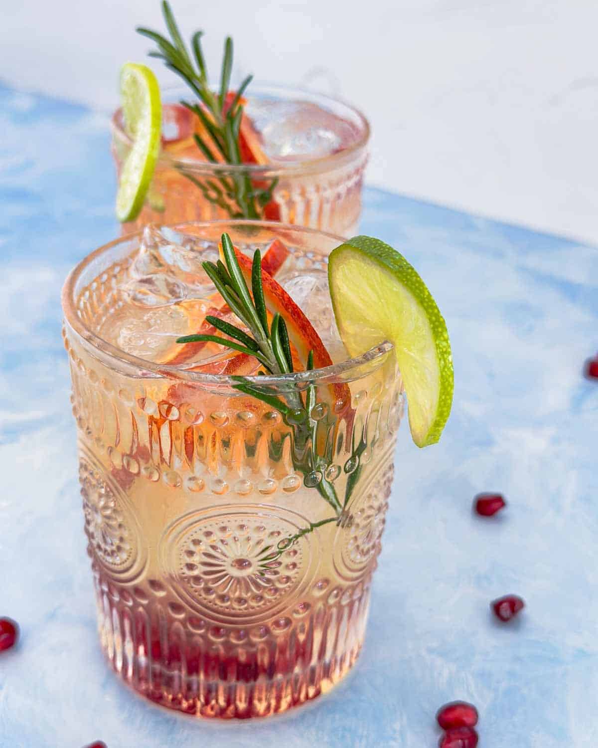 Side view of glasses of white wine sangria on a blue background showing lime slices, rosemary sprigs, and pomegranate seeds.