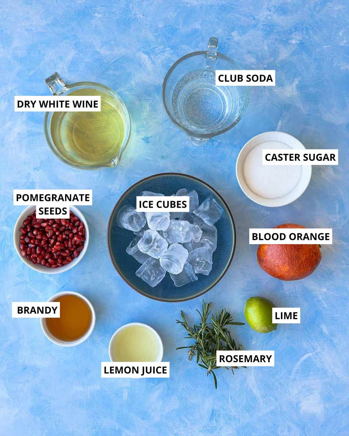 Labeled overhead image of white sangria ingredients arranged on a blue background, including dry white wine, club soda, caster sugar, pomegranate seeds, brandy, lemon juice, ice cubes, blood orange, lime, and fresh rosemary.