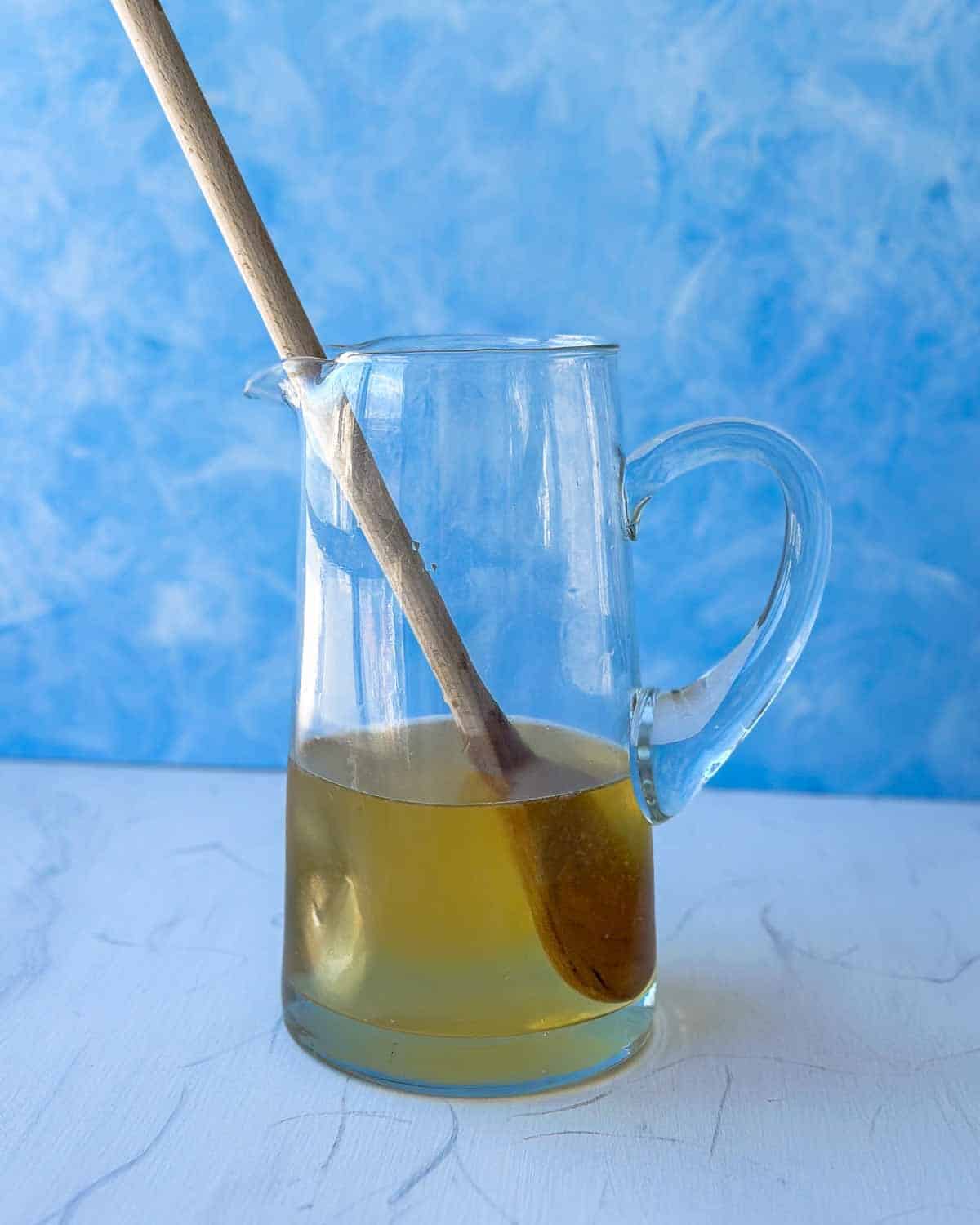Pitcher filled with dry white wine being stirred with a wooden spoon against a blue and white backdrop, the first step in making homemade white sangria.
