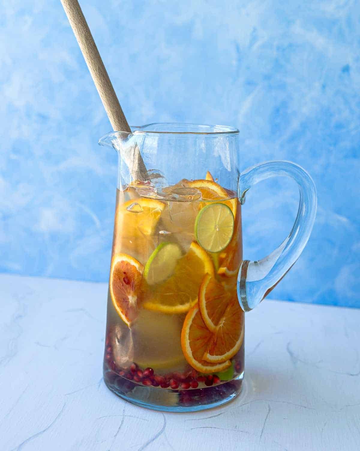 Pitcher of white sangria with sliced blood orange, lime, and pomegranate seeds being stirred with a wooden spoon, showing the fruit-infusing step of the recipe.