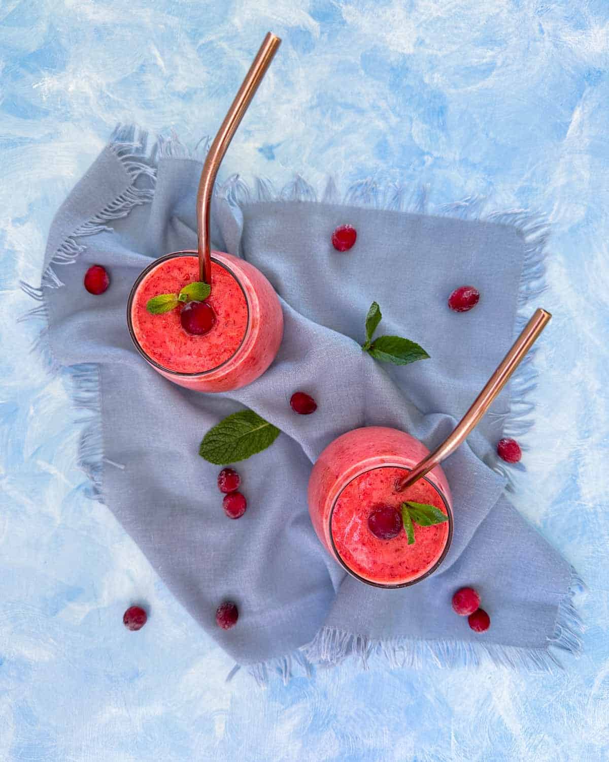 Overhead view of cranberry smoothies arranged on a soft blue cloth, styled with fresh cranberries and mint for a colorful, seasonal look.