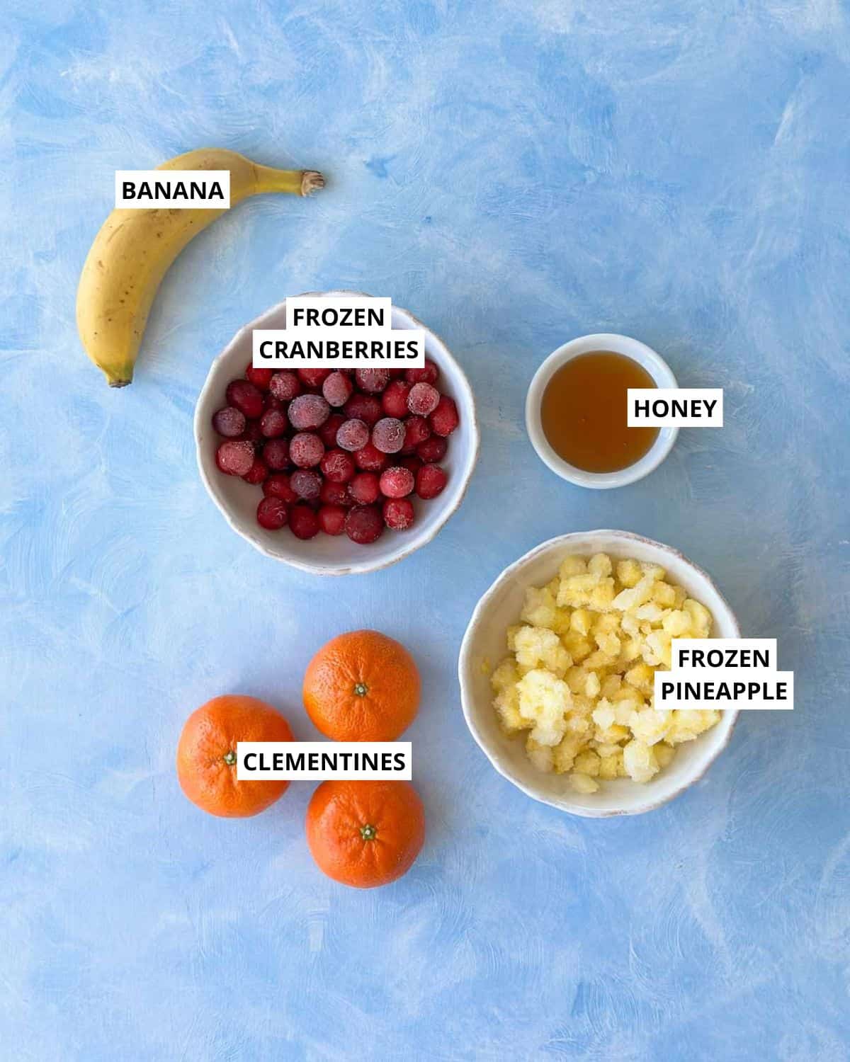 Overhead view of ingredients for a cranberry smoothie, including a ripe banana, frozen cranberries, frozen pineapple, clementines, and honey arranged on a soft blue background.