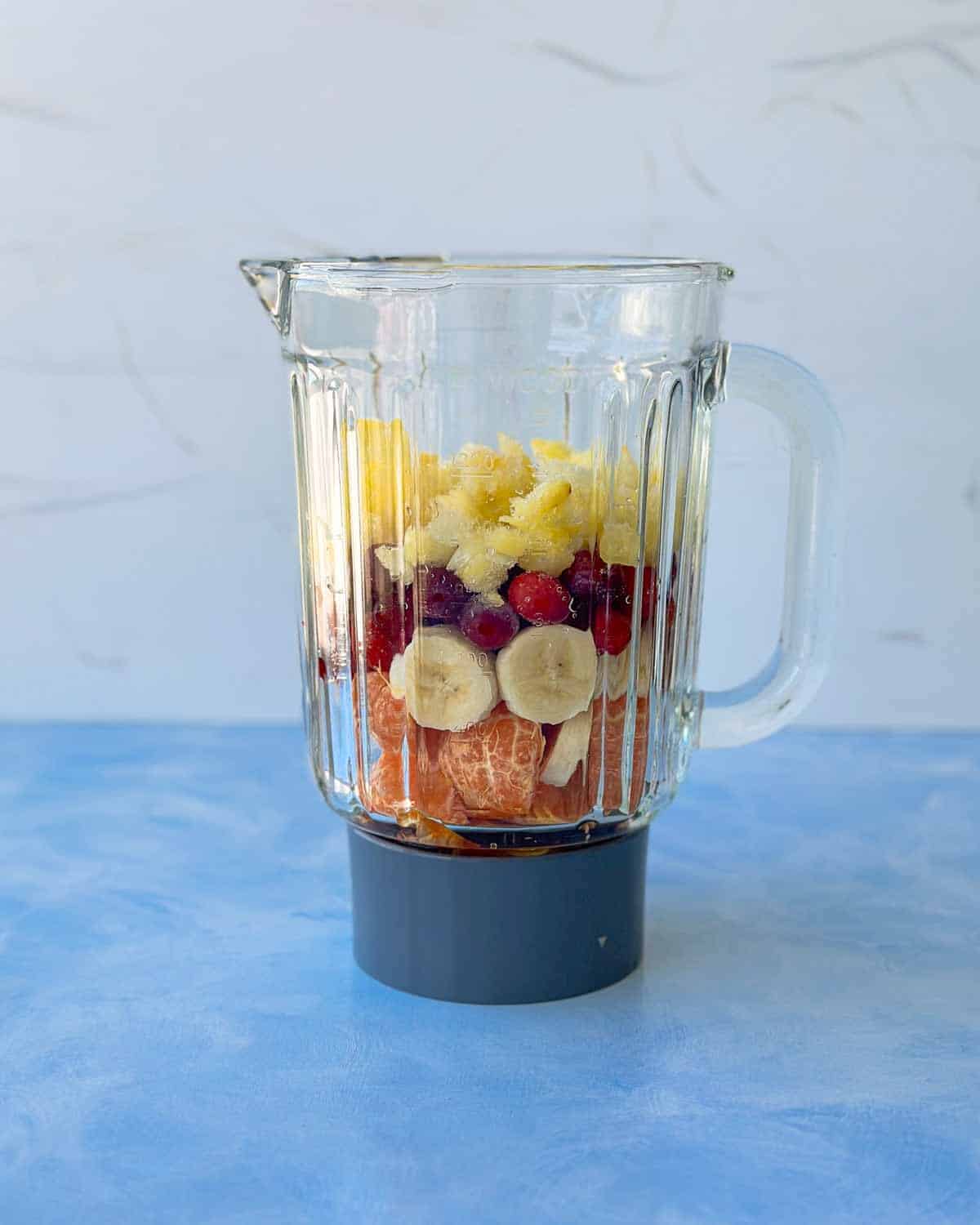 Blender filled with layered cranberry smoothie ingredients — banana slices, clementine segments, frozen cranberries, pineapple, and honey — ready to blend.
