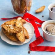 Close-up of golden homemade Italian almond biscotti on a plate with coffee and festive red ribbon details in the background.