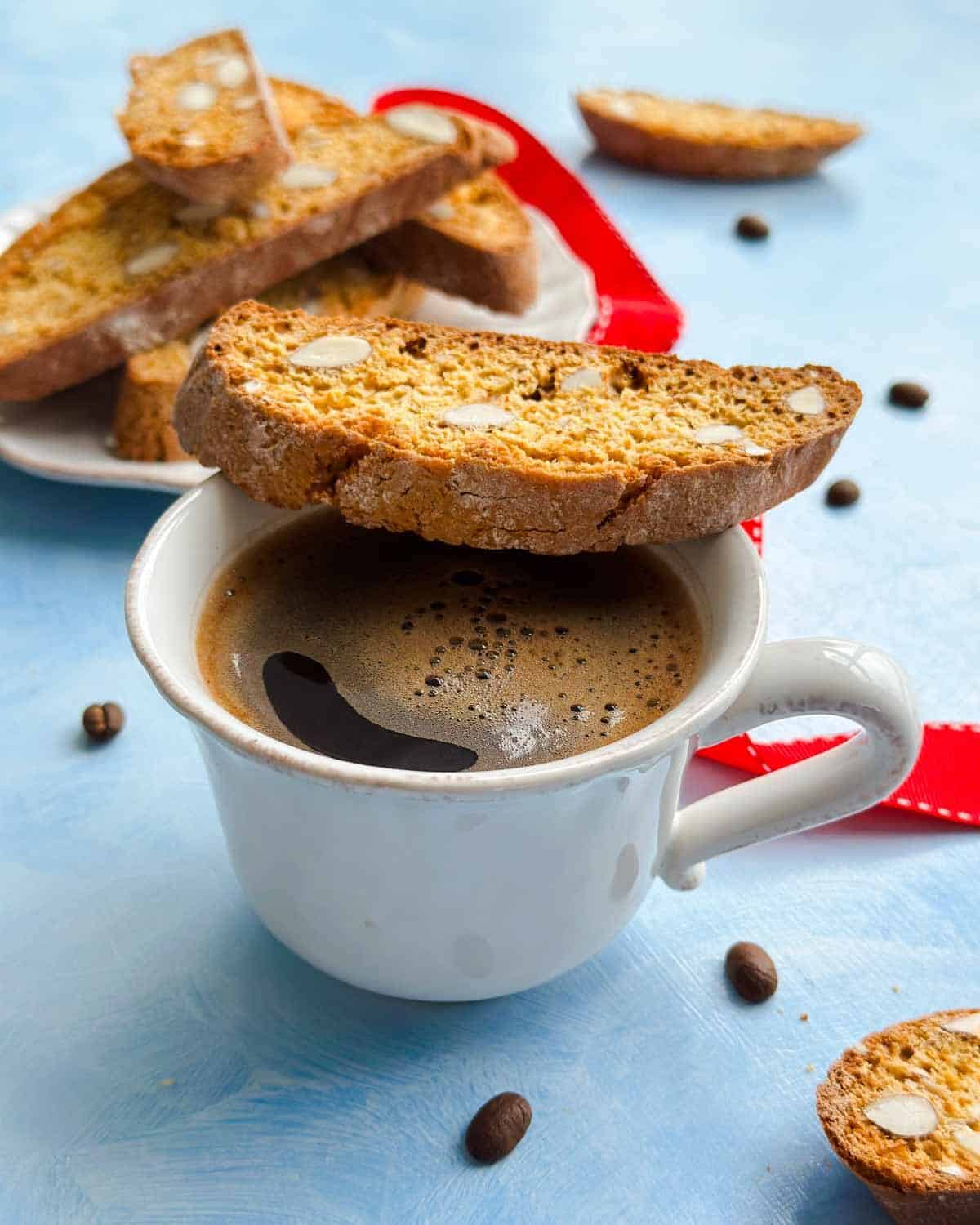 Side-angle close-up showing crisp almond biscotti stacked on a plate with coffee cups and scattered coffee beans.