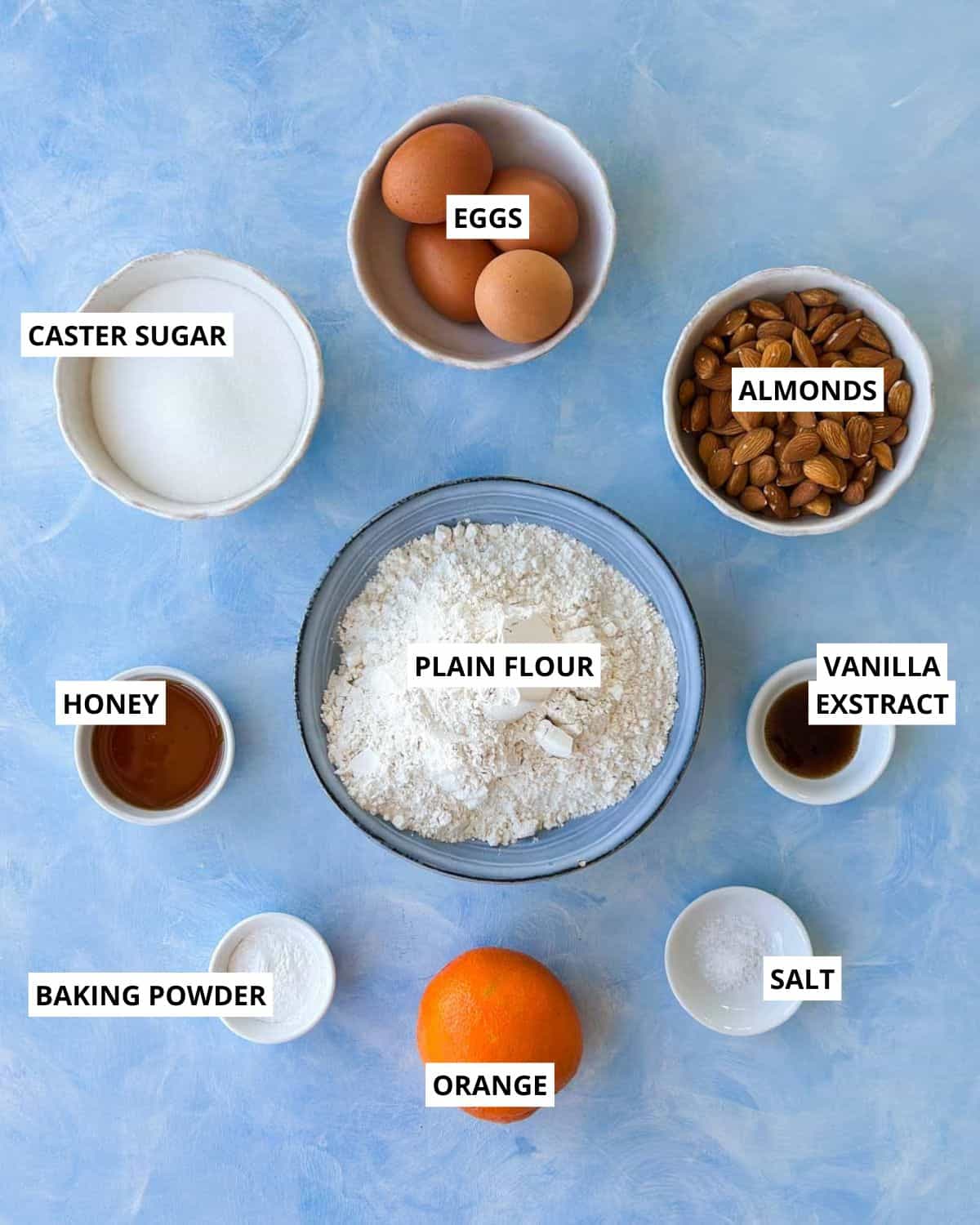 Overhead shot of ingredients for Italian almond biscotti, including plain flour, caster sugar, whole almonds, eggs, honey, vanilla extract, baking powder, salt, and an orange, arranged neatly on a light blue background.