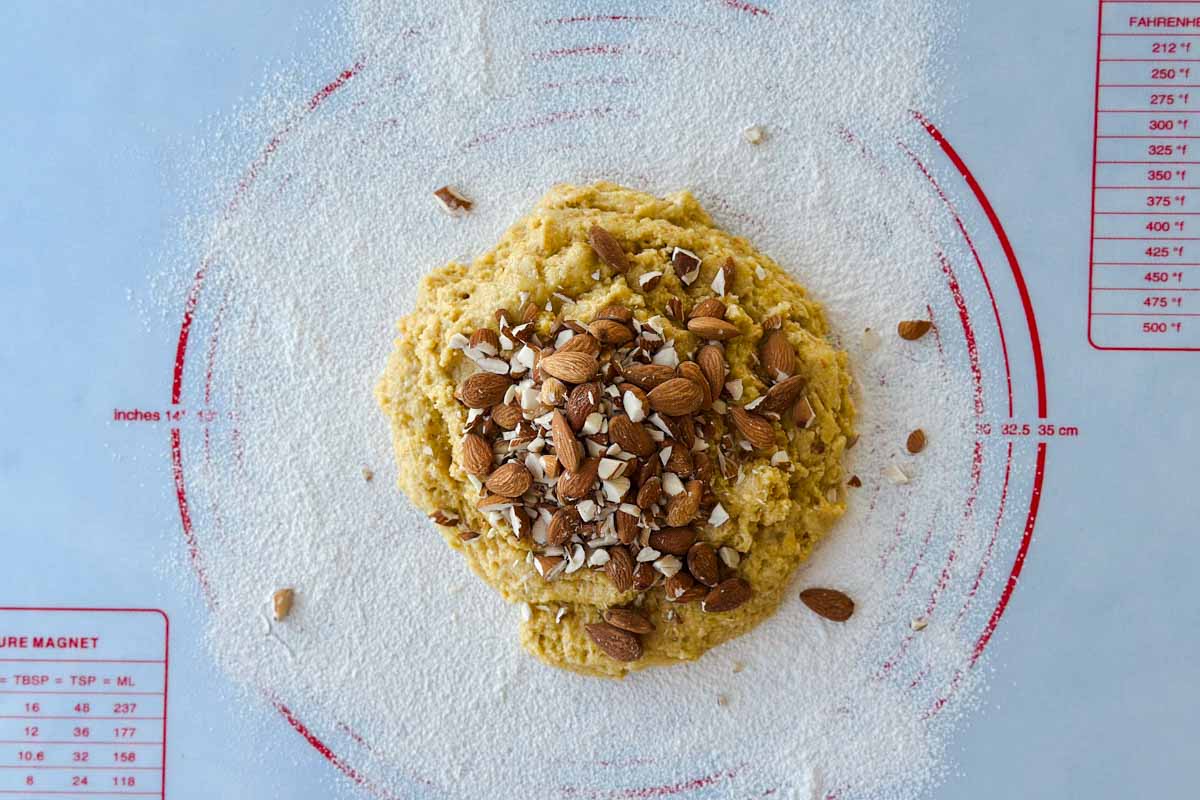 Almond biscotti dough tipped onto a floured mat and topped with whole almonds, ready to be kneaded.
