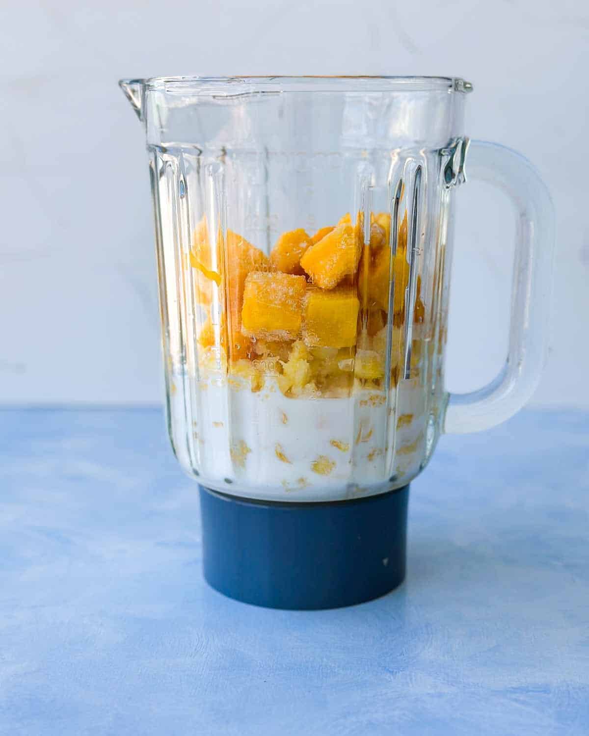 Blender jug filled with frozen mango, pineapple, banana and almond milk ready to be blended into a smoothie.